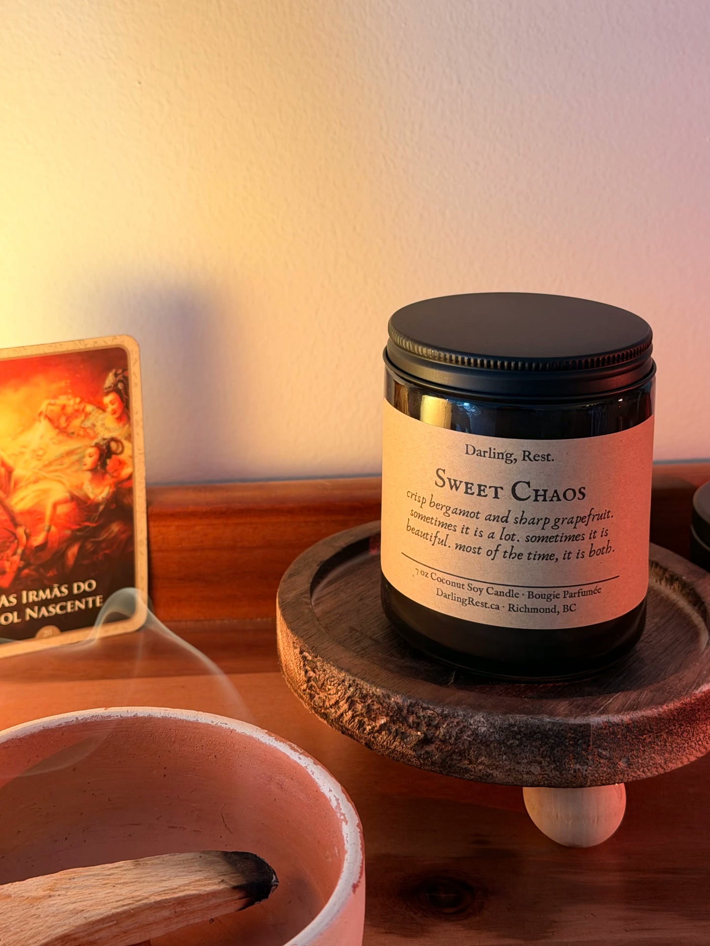 Amber Sweet Chaos candle with a black lid on a rustic wooden riser, a smoldering palo santo bowl in the foreground and an oracle card to the left in warm golden light