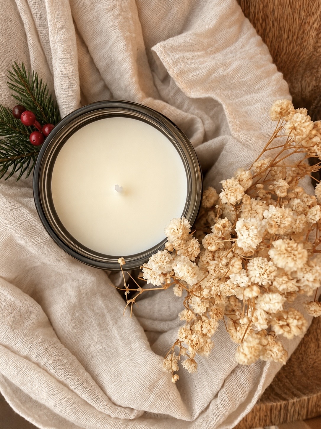 Overhead view of an unlit cream candle in a dark glass jar on draped linen, a pine sprig with red berries at the upper left and dried baby's breath at the lower right